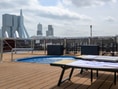 Deck eines Schiffes mit Pool und Liegestühlen, im Hintergrund eine Brücke und Wolkenkratzer.