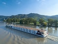 Kreuzfahrtschiff auf Fluss vor bewaldeten Hügeln.