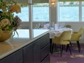 Innenansicht eines eleganten Restaurants mit Blumenarrangement, Tisch und Stühlen in der Nähe eines grossen Fensters mit Blick auf das Wasser.