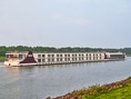 Langes Kreuzfahrtschiff auf einem Fluss mit grünem Ufer.