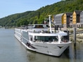 Flusskreuzfahrtschiff an einem Flussufer mit Hügeln und Gebäuden im Hintergrund.