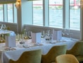 Eleganter Restaurantbereich mit gedeckten Tischen und Flussblick durch grosse Fenster.