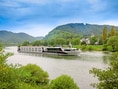 Flusskreuzfahrtschiff auf dem Fluss vor bewaldeten Hügeln.