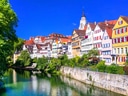 Bunte Altstadthäuser am Flussufer mit Kirchturm im Hintergrund.