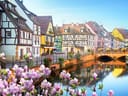 Malerische Fachwerkhäuser am Fluss mit Brücke und Blumen im Vordergrund.