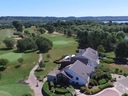 Luftaufnahme eines Golfplatzes mit Clubhaus, Bäumen und See im Hintergrund.
