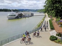 Radfahrer auf einem Weg entlang eines Flusses mit anlegendem Schiff.