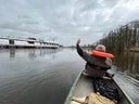 Person in einem Kanu winkt einem vorbeifahrenden Schiff zu, auf einem ruhigen Fluss.