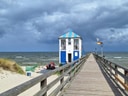 Steg am Strand mit kleinem blau-weissem Gebäude und Wolkenhimmel.