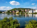 Flussufer mit Gebäuden im Hintergrund und bewaldetem Hügel.