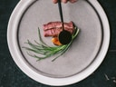 Ein Teller mit Fleisch, Gemüse und eine Hand mit einem Löffel Sosse.