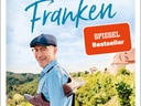 Ein Mann mit Mütze steht draussen vor einer hügeligen Landschaft.