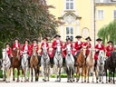 Gruppe von Reitern in roten Jacken auf einer Allee vor einem Gebäude.