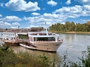 Flusskreuzfahrtschiff auf ruhigem Fluss mit Bäumen und blauem Himmel.