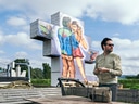 Mann mit Sonnenbrille vor grossem Wandgemälde im Freien, mit Wolkenhimmel.