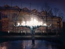 Leuchtende Skulptur eines Roboters mit ausgebreiteten Armen vor einem Gebäude, spiegelt sich im Wasser.