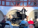 Musiker im Frack singt neben Pianistin auf Innenhofbühne vor Publikum.