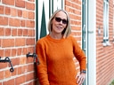 Frau mit Sonnenbrille und orangefarbenem Pullover vor Backsteinmauer.