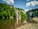 Flussufer mit Kieselsteinen und bewaldeten Kalksteinfelsen im Hintergrund.