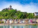 Burg auf bewaldetem Hügel über bunten Fachwerkhäusern am Fluss.