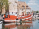 Rotes Fischerboot im Hafen neben einem älteren Gebäude, Menschen gehen am Ufer entlang.