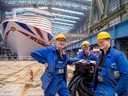 Drei Arbeiter in blauer Kleidung und gelben Helmen vor einem grossen Schiff im Trockendock.