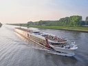 Flusskreuzfahrtschiff auf einem breiten Fluss mit grünen Ufern.