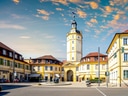 Städtischer Platz mit historischem Turm und umliegenden Gebäuden bei Sonnenuntergang.