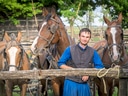 Mann in traditioneller Kleidung steht mit drei braunen Pferden an einem Holzzaun.