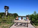 Drei Personen auf einer Bank vor Aussichtsturm und See.