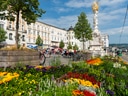 Bunte Blumenbeete und barocke Denkmäler auf einem belebten städtischen Platz.