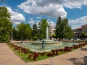 Springbrunnen in einem Park mit Bäumen und Gebäuden im Hintergrund.