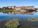 Gebäude am Ufer eines ruhigen Flusses mit Seerosen im Vordergrund.