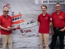 Drei Personen in roten Hemden stehen vor einem Bild eines Rettungsboots.