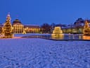 Verschneiter Platz mit beleuchteten Bäumen und einem Brunnen vor einem grossen Gebäude am Abend.