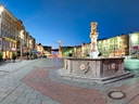 Beleuchteter Platz mit Brunnen und historischen Gebäuden bei Dämmerung.
