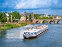 Flusskreuzfahrtschiff auf einem Fluss mit Brücke und Altstadt im Hintergrund.