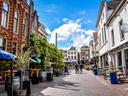 Strassenszene mit Menschen zwischen historischen Gebäuden und Pflanzen, blauer Himmel.