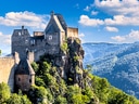 Burg auf einem Hügel mit Wald und Bergen im Hintergrund.
