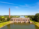 Altes Industriegebäude mit Schornstein am Wasser, umgeben von Landschaft.