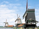 Drei Windmühlen an einem Flussufer unter blauem Himmel.