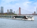 Flusskreuzfahrtschiff auf dem Wasser vor einer modernen Stadtbrücke mit Hochhäusern im Hintergrund.