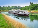 Flusskreuzfahrtschiff auf einem ruhigen Kanal mit grünen Ufern.