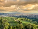 Panoramablick auf eine Stadt mit grünen Hügeln und dramatischem Himmel.