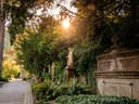 Friedhofsszene mit Grabmal, Statue und Bäumen bei Sonnenuntergang.