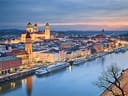 Panoramablick auf eine Stadt an einem Fluss bei Sonnenuntergang, mit beleuchteten Gebäuden und Kirchen.