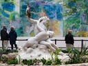 Weisse Skulptur mit umarmenden Figuren in einem Garten, Gemälde im Hintergrund.