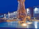 Historischer Stadtplatz bei Nacht mit Brunnen im Vordergrund und Gebäuden im Hintergrund.