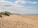 Weitläufiger Sandstrand mit Gebäuden und Dünen im Hintergrund.