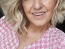 Frau mit blonder, welliger Frisur und rosa-weissem Mantel, lächelt.